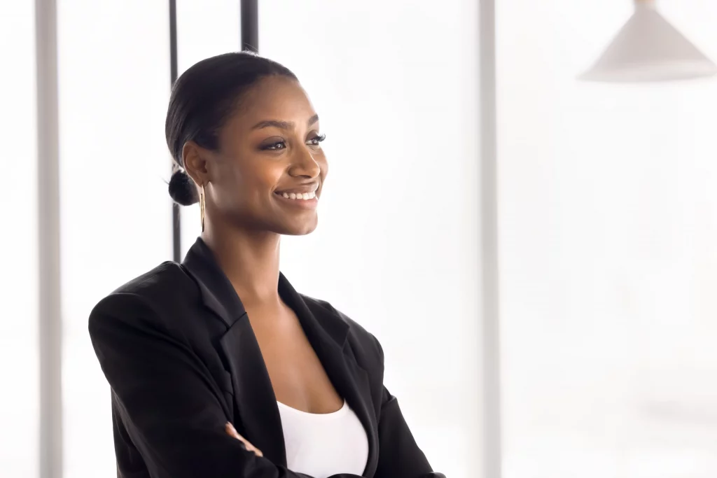 Femme souriante en blazer noir, bras croisés, dans un bureau lumineux
